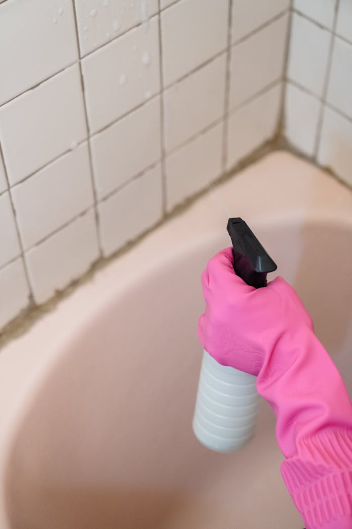 services-01 A person wearing a pink glove cleans bathroom tiles with a spray bottle.